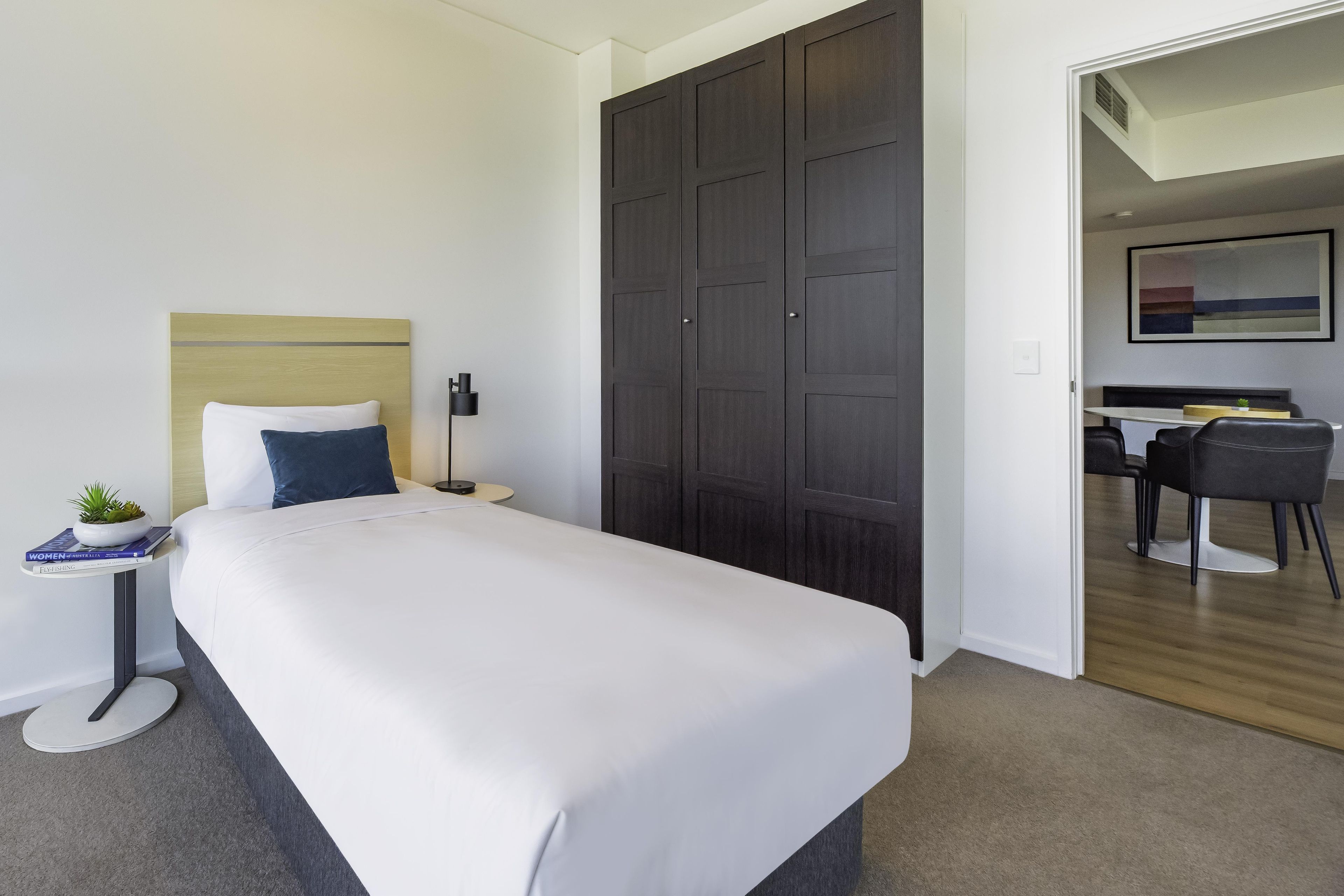Quarto de hotel com cama, mesa de cabeceira, abajur e um guarda-roupa escuro.