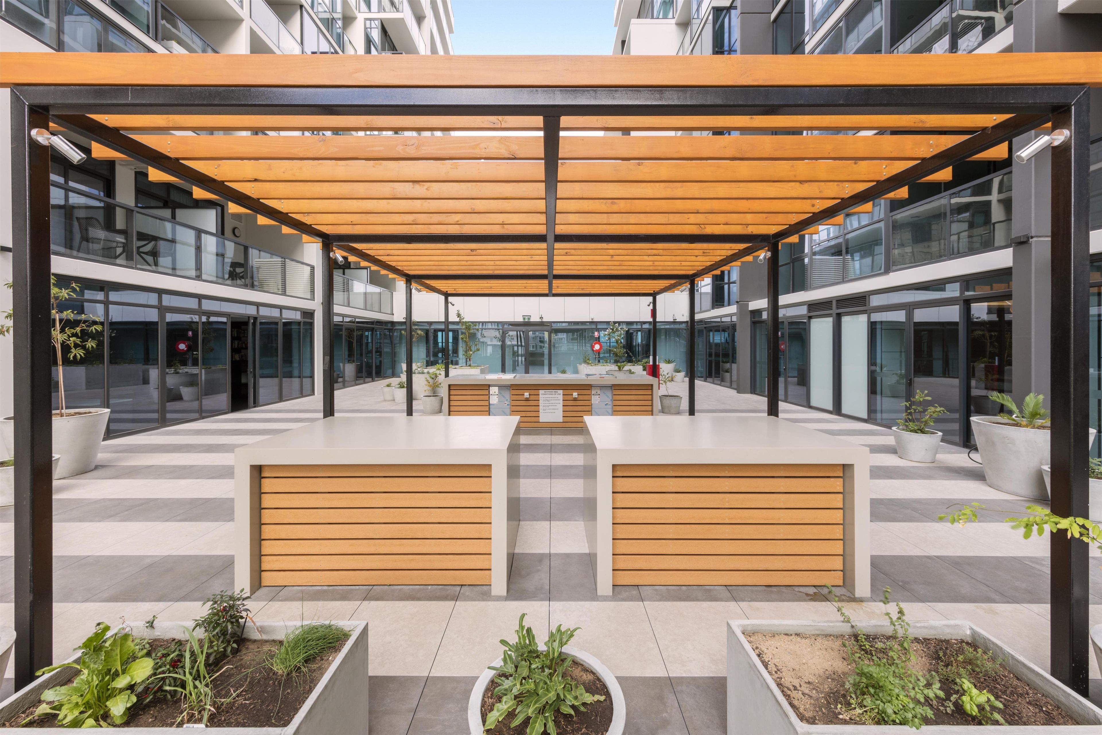 Un área de estar exterior moderna con bancos de madera y macetas, rodeada por paredes de vidrio y una barandilla de metal.