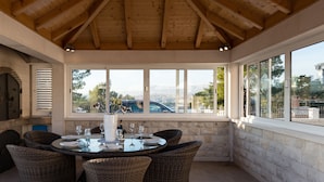 A covered patio with a round table and wicker chairs, large windows, and a stone wall. - Villa Miljak (Podstrana)