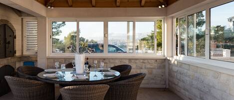 A covered patio with a round table and wicker chairs, large windows, and a stone wall.