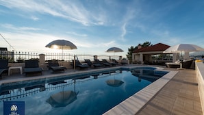 A swimming pool with lounge chairs and umbrellas, surrounded by a fence and a building with a red roof. - Villa Miljak (Podstrana)