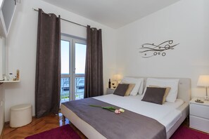 A modern hotel room with a large bed, a nightstand, a window with curtains, and a view of the outdoors. - Ante apartments (Podstrana)