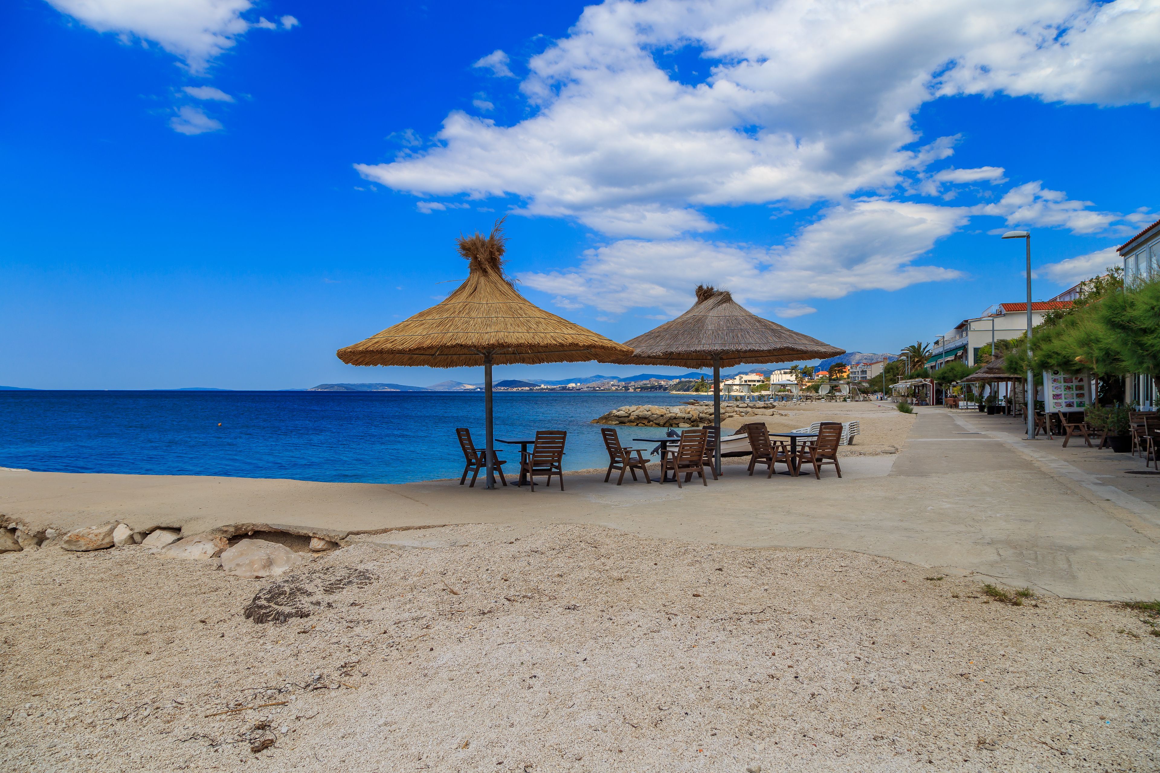 A beachside café with tables and chairs under thatched umbrellas, overlooking the sea.