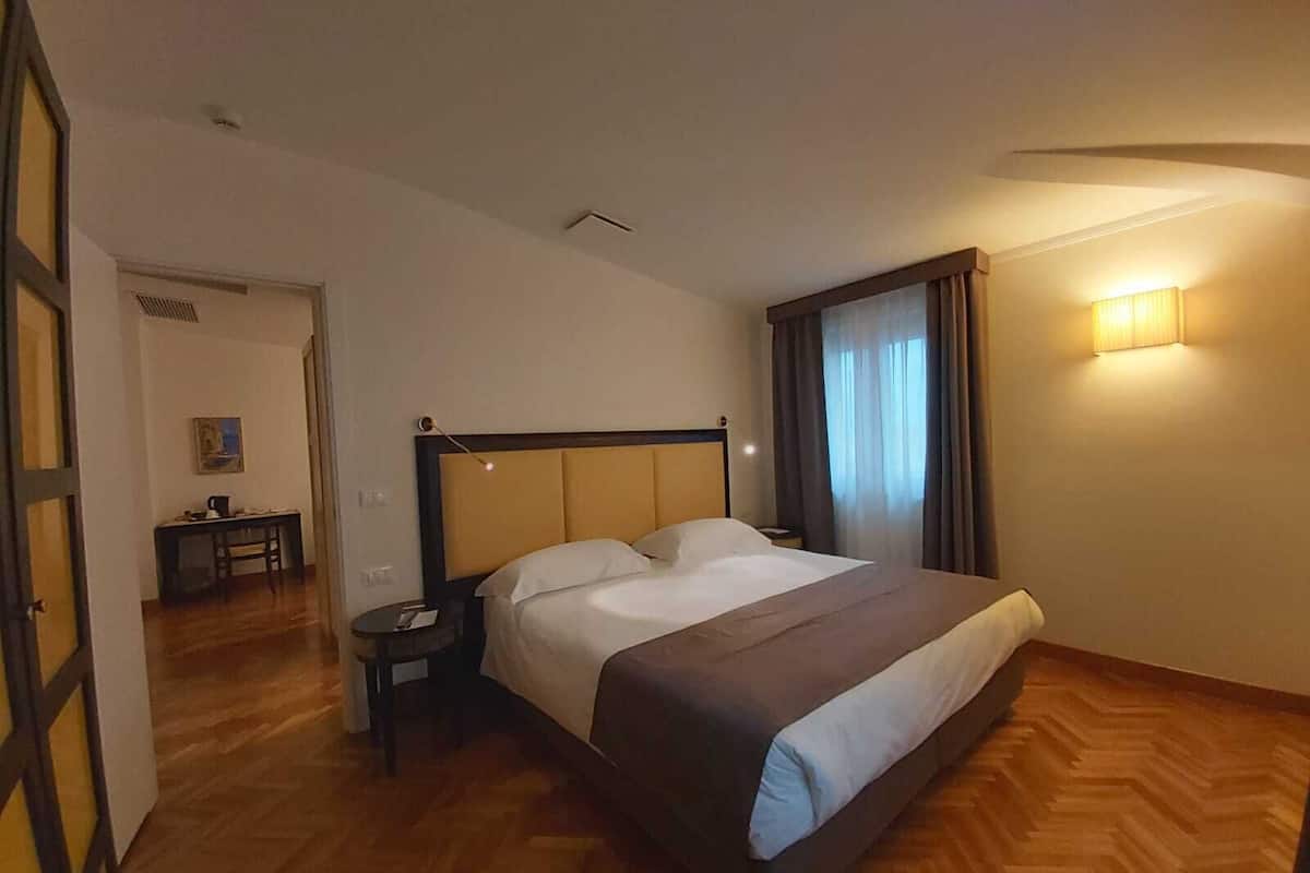 A hotel room with a large bed, a bedside table, a lamp, a chair, and a desk with a painting.