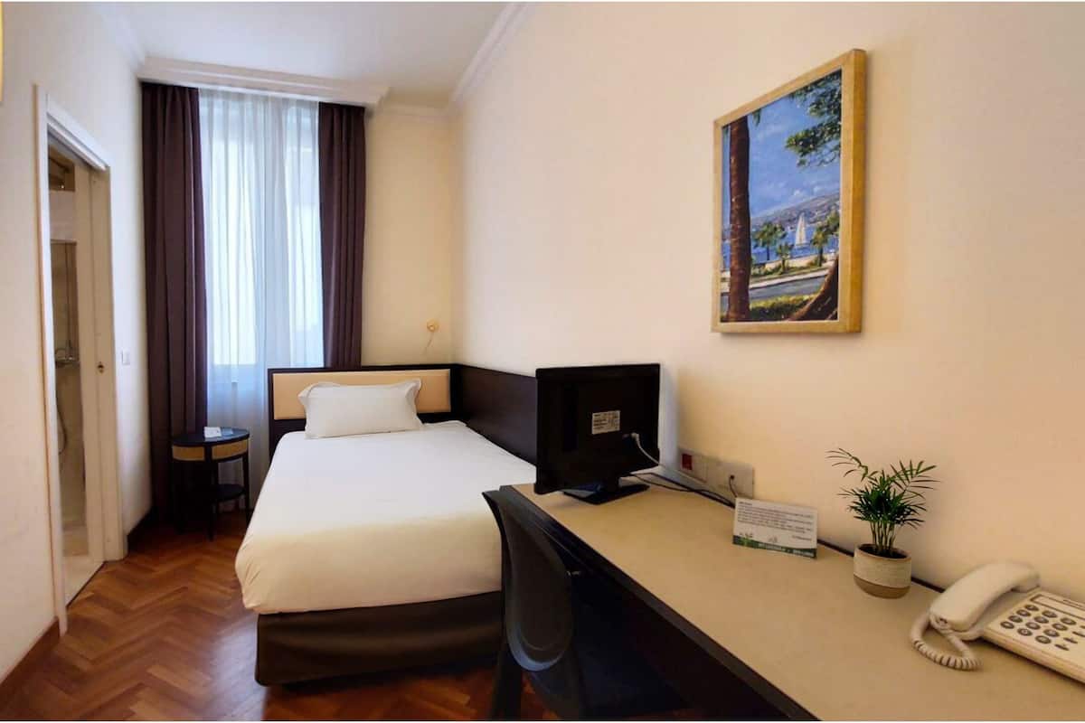 A hotel room with a bed, desk, computer, phone, and a painting on the wall.