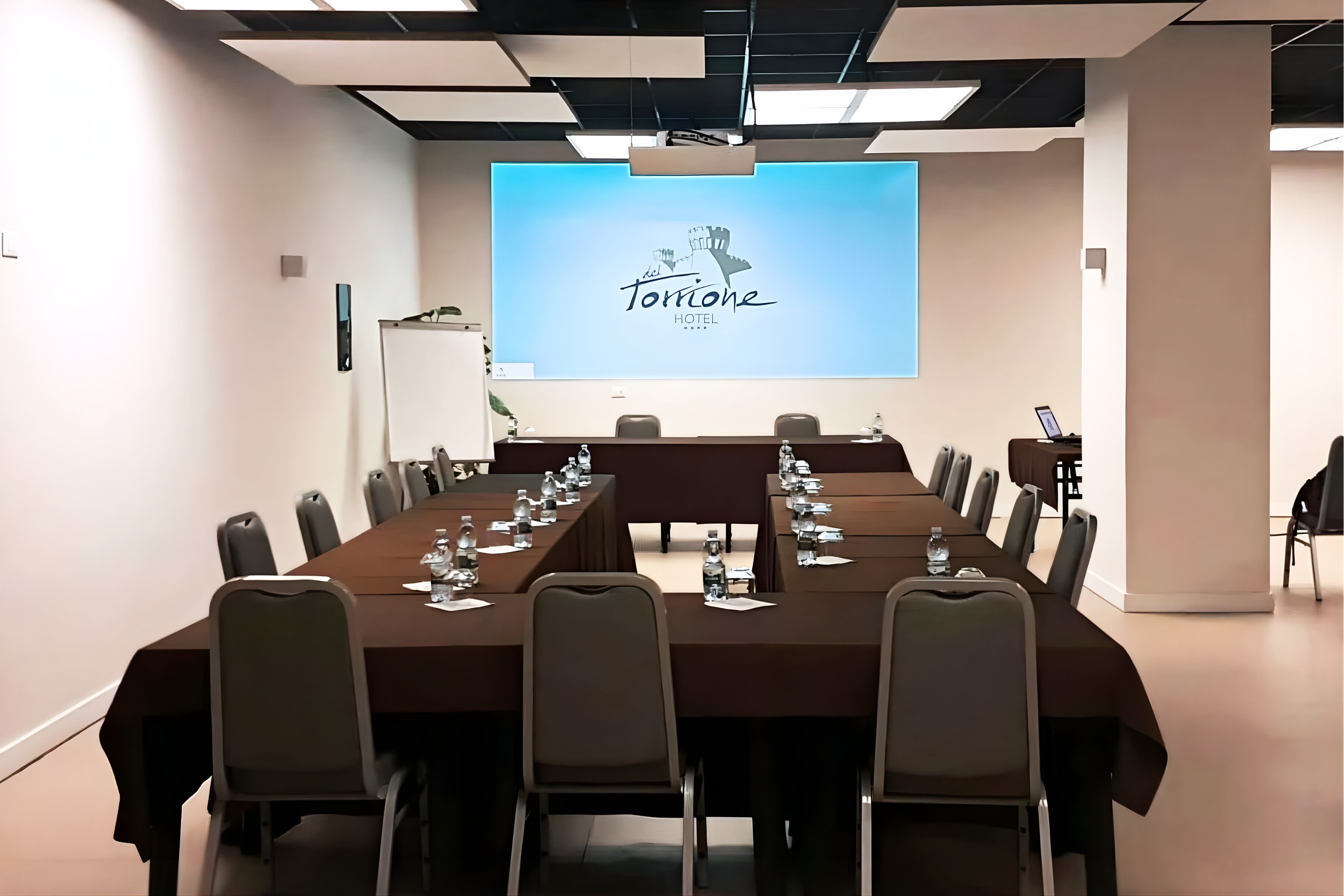 A conference room with a long table, chairs, a projector screen displaying 'Torione Hotel,' and a whiteboard.