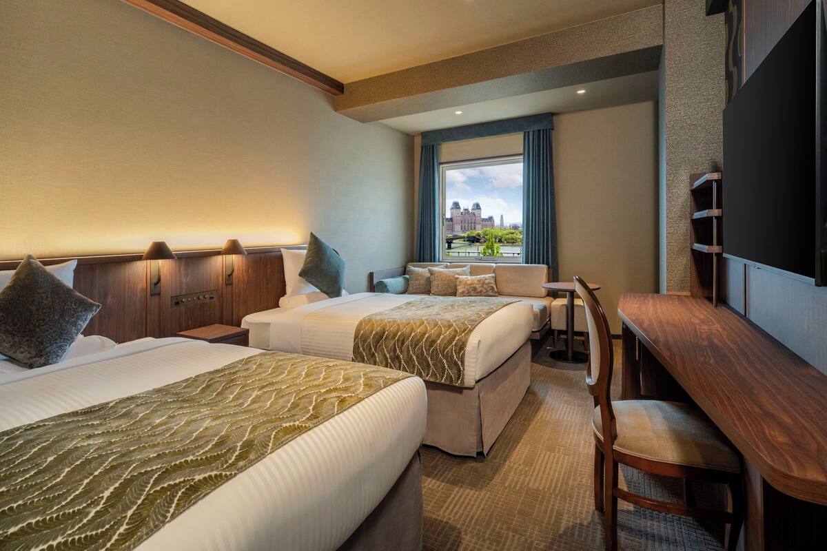 A hotel room with two beds, a desk, and a large window offering a city view.