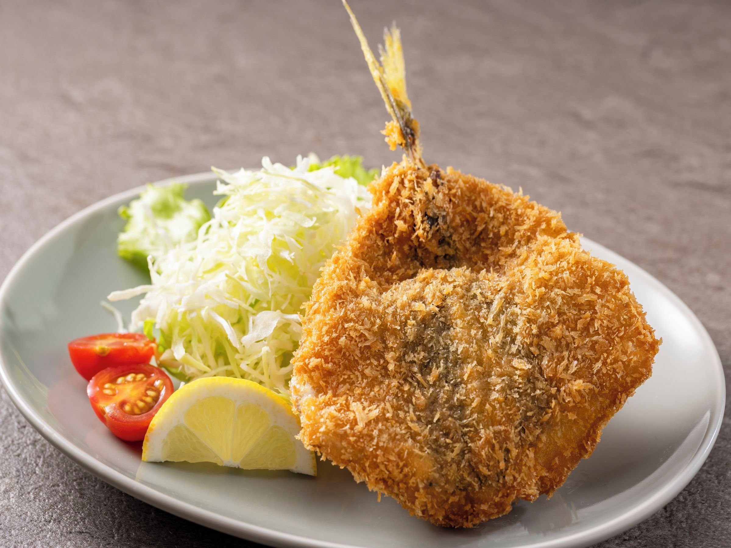 A plate with a breaded and fried item, possibly a fish fillet, accompanied by a side of shredded cabbage, a slice of lemon, and a cherry tomato.