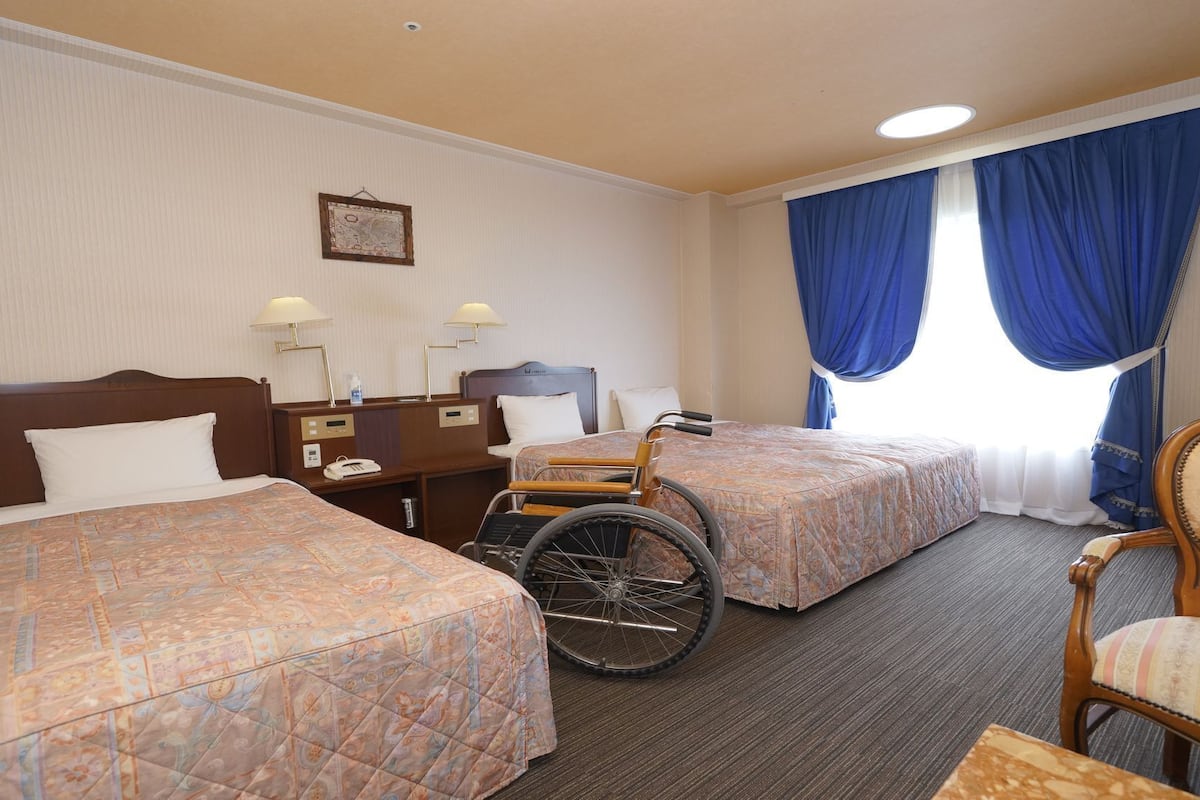 A hotel room with two beds, a wheelchair, a chair, a nightstand, and a bedside table with lamps.
