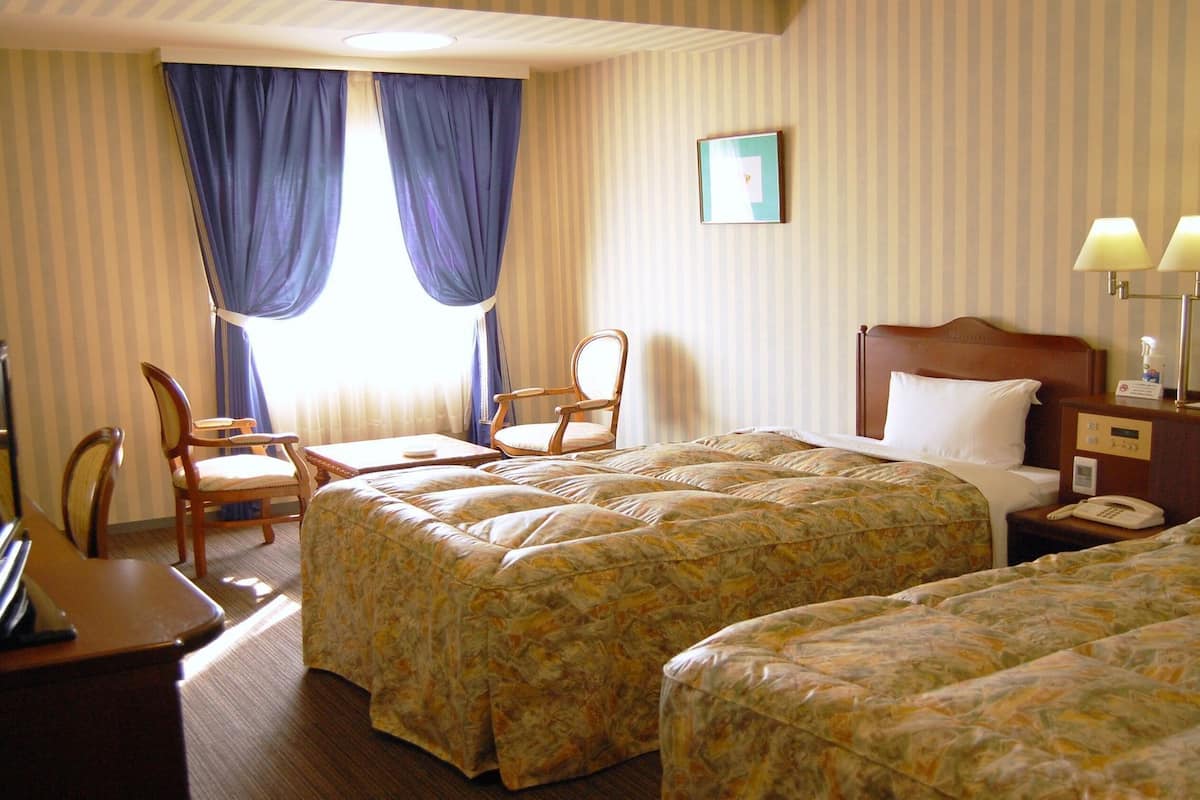 A hotel room with two beds, a desk with a television, a chair, and a bedside table with lamps.