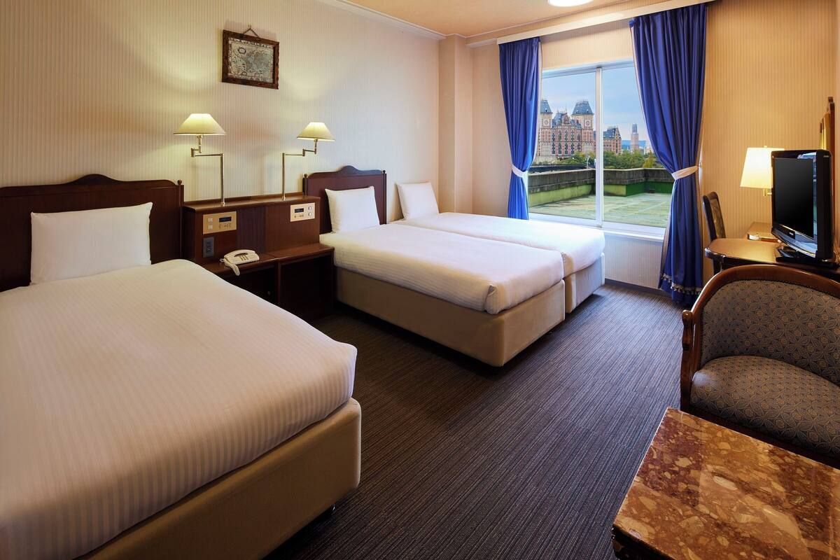 A hotel room with two beds, a desk, a chair, a TV, and a window with curtains.