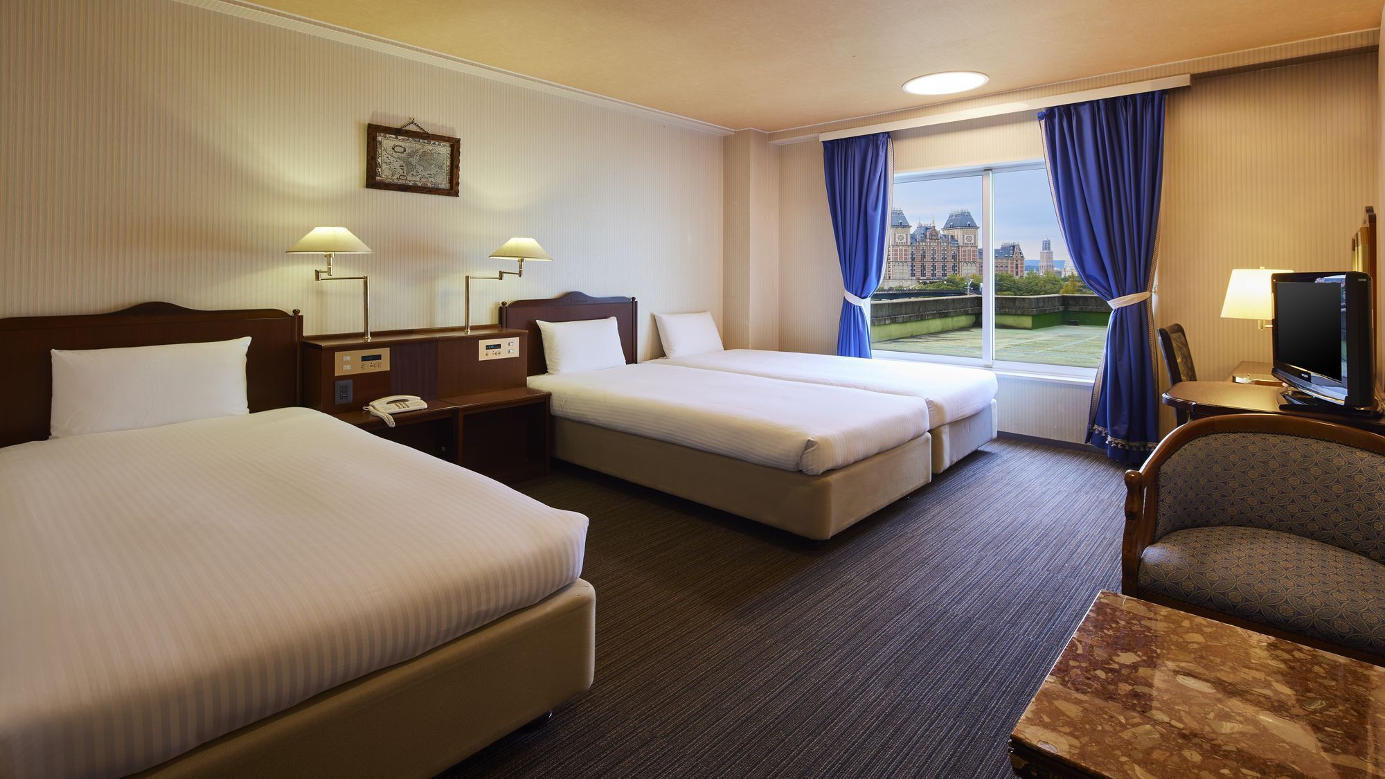 A hotel room with two beds, a desk, a chair, a television, and a window with a view of buildings.