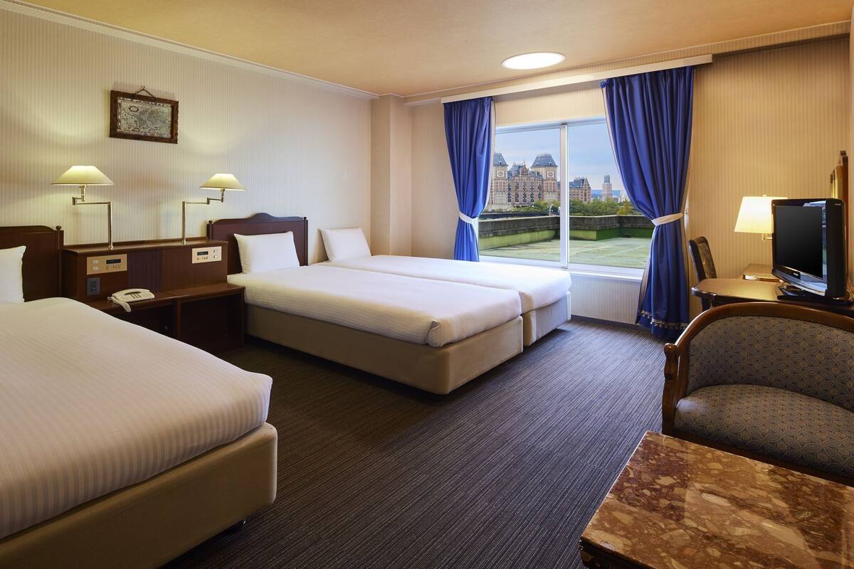 Habitación de hotel con dos camas, un escritorio, una silla, un televisor y una ventana con vistas a edificios.