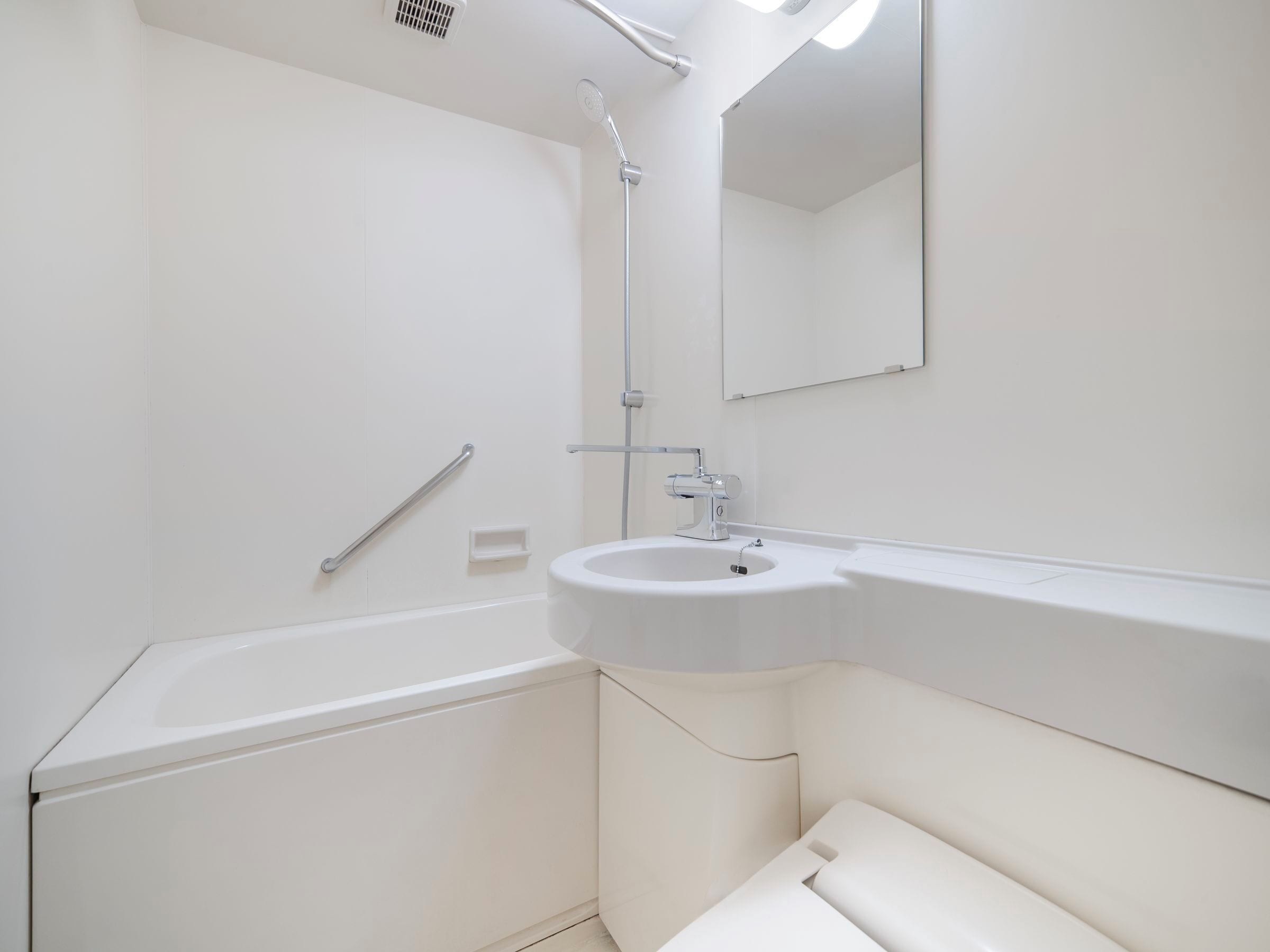 A bathroom with a white bathtub, a sink, a showerhead, a handrail, and a mirror.