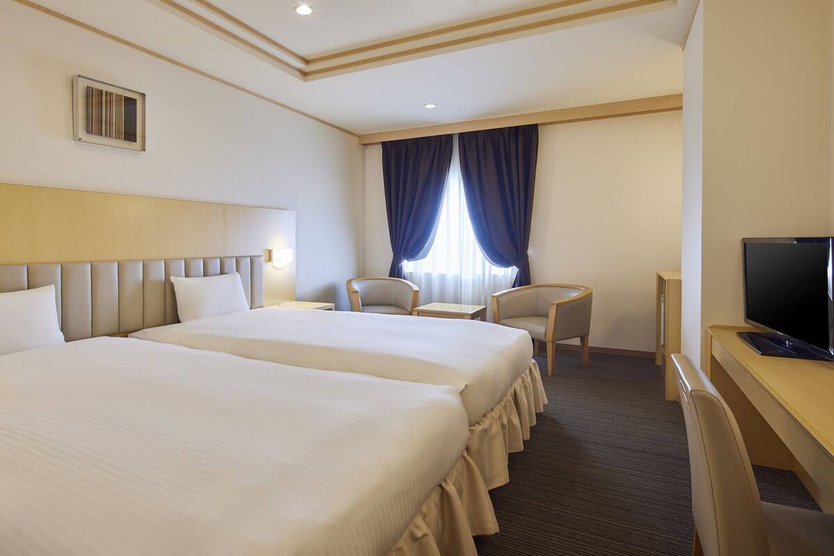 Habitación de hotel con dos camas, un escritorio, una silla, un televisor y una ventana con cortinas.