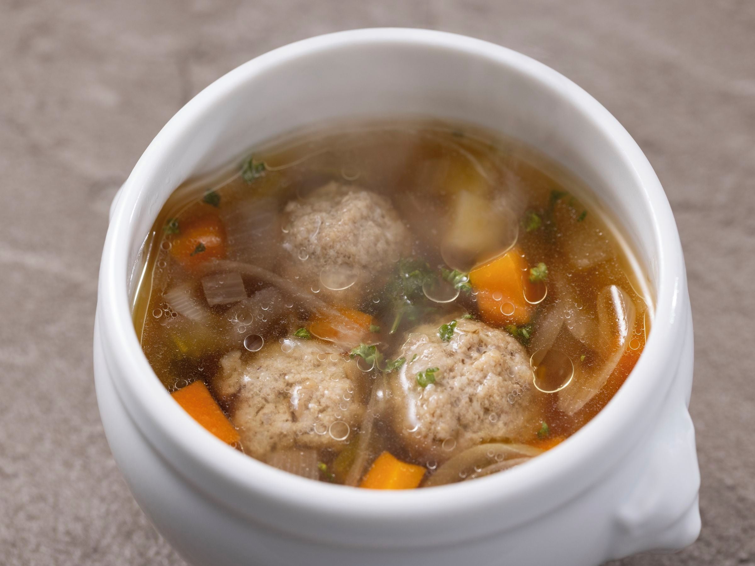 Un tazón de cerámica blanca con caldo claro, albóndigas, zanahorias y hierbas.