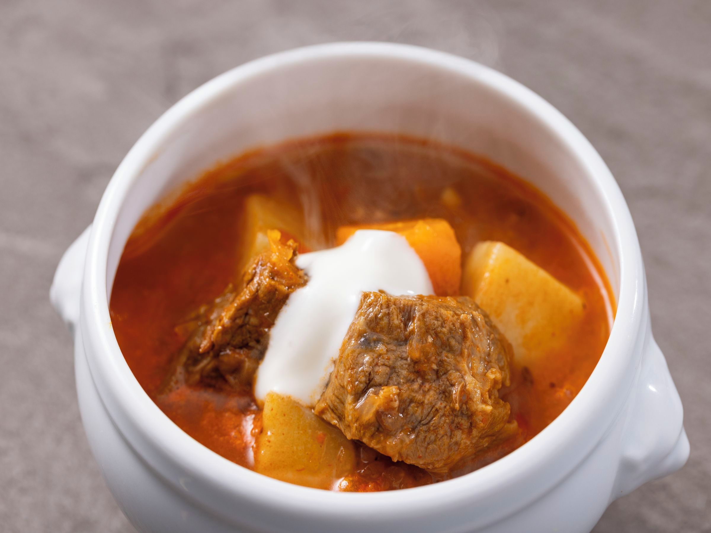 Un tazón de cerámica blanca con un guiso de carne y papas, coronado con una cucharada de crema.