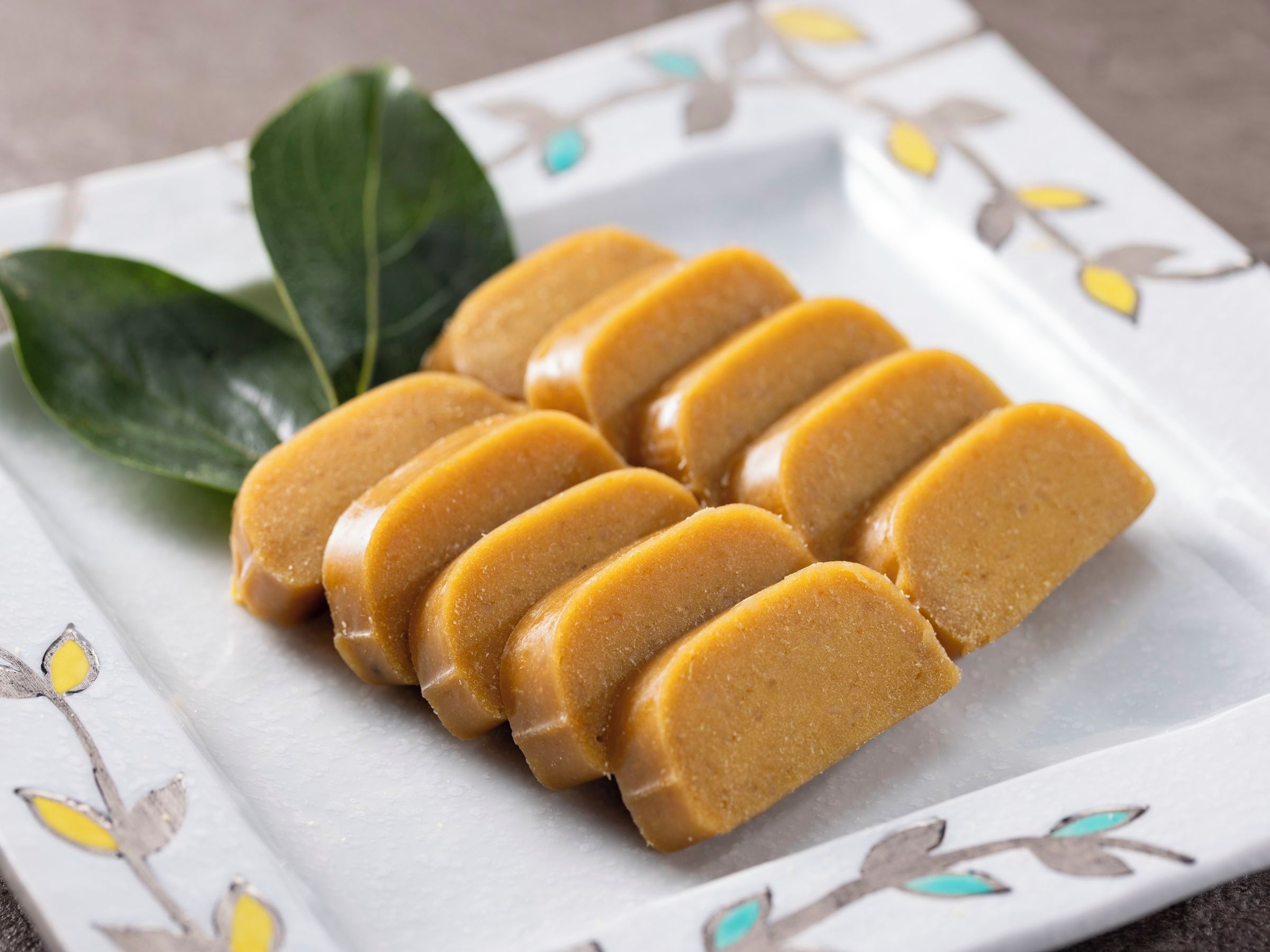 Un plato blanco con estampado floral, que contiene varias porciones de un alimento amarillo, posiblemente un postre, acompañado de hojas verdes.