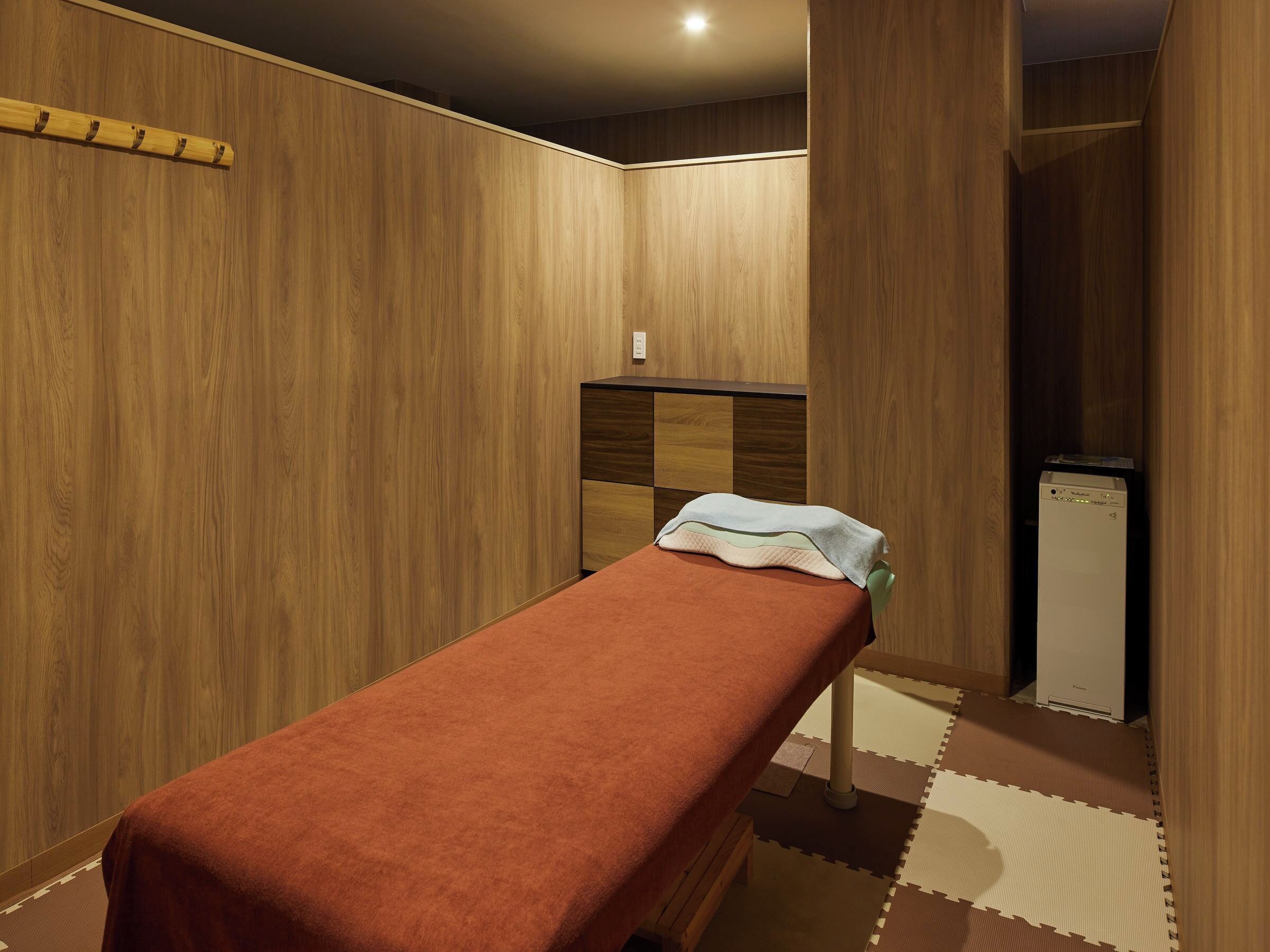 A room with a wooden wall, a bed with a red blanket, a small cabinet, and a refrigerator.