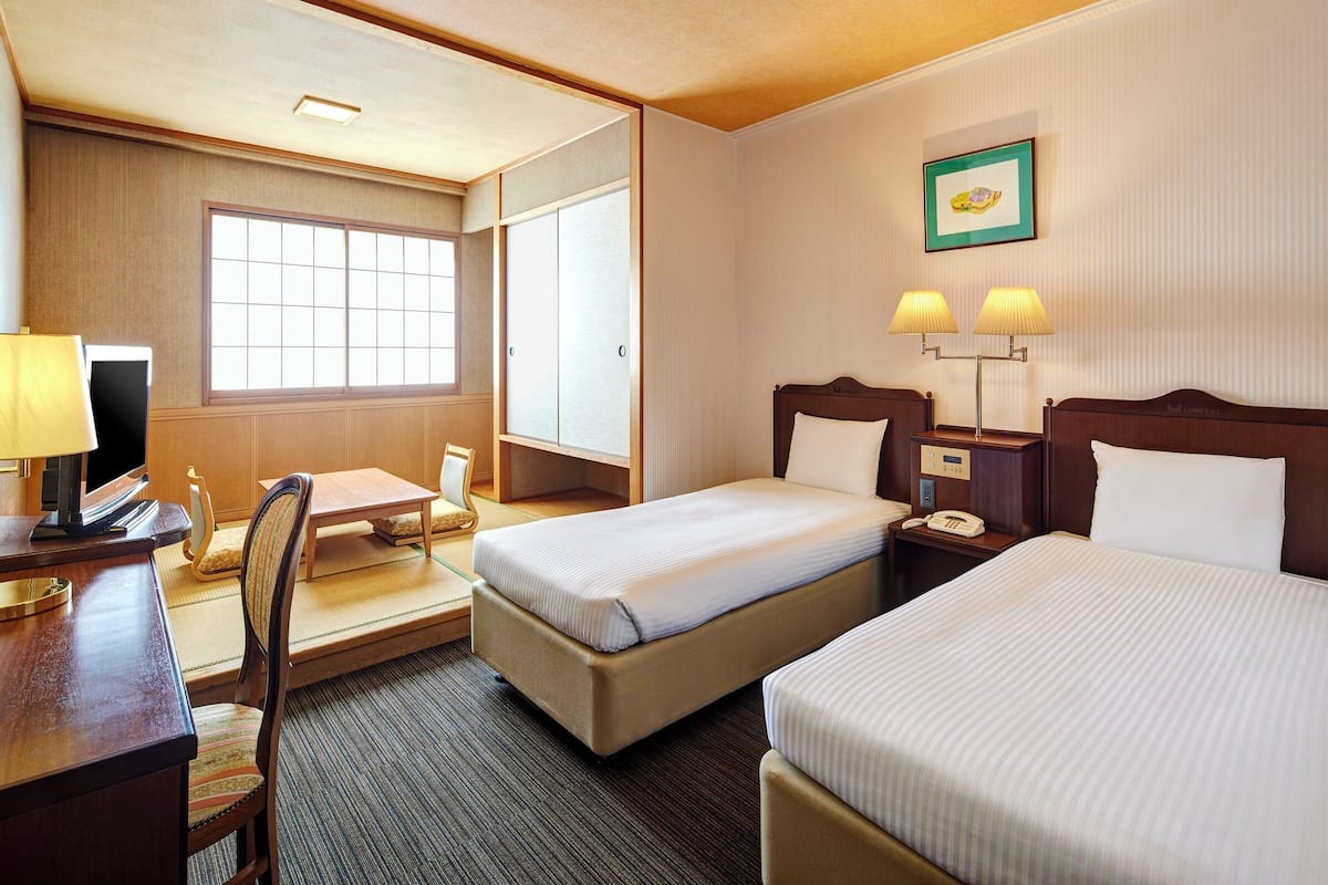 A hotel room with two beds, a desk with a computer, a chair, and a window.