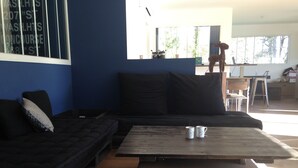Una sala de estar moderna con un sofá de color oscuro, una mesa de centro de madera con dos tazas, y un ventanal grande que proporciona luz natural.