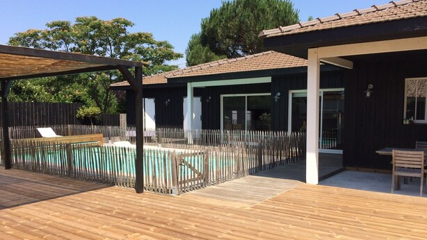 Una terraza de madera con piscina, un porche cubierto y un edificio con grandes ventanales.