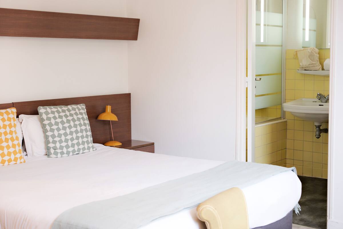 Une chambre d’hôtel avec un lit, une table de chevet, une lampe et une salle de bain visible à travers une porte ouverte.