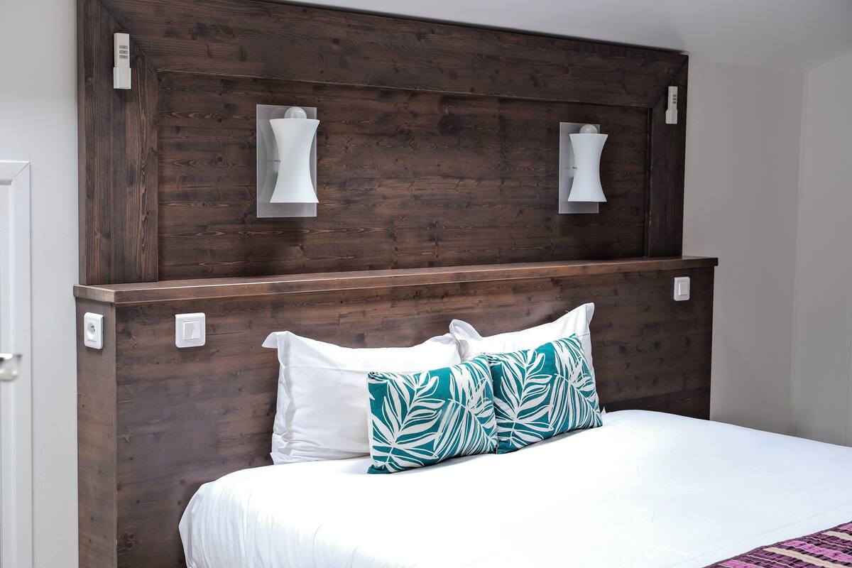 Une chambre avec une tête de lit en bois, une literie blanche et un coussin à motifs de feuilles turquoise.