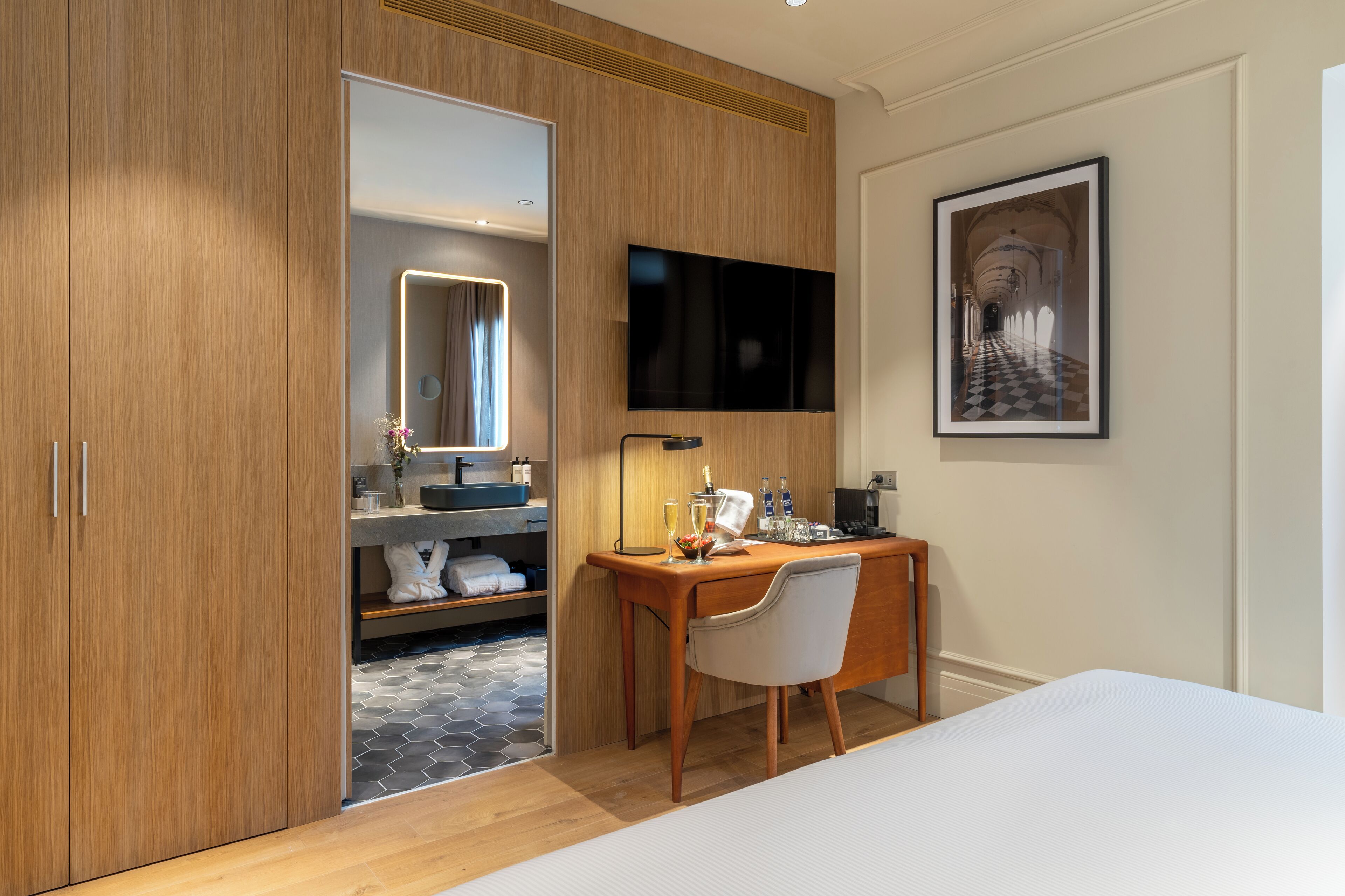 A modern hotel room with a wooden wardrobe, a desk with a chair, a flat-screen TV, a framed picture on the wall, and a bed.