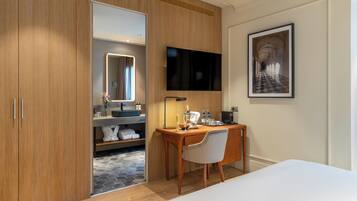 A modern hotel room with a wooden wardrobe, a desk with a chair, a flat-screen TV, a framed picture on the wall, and a bed.