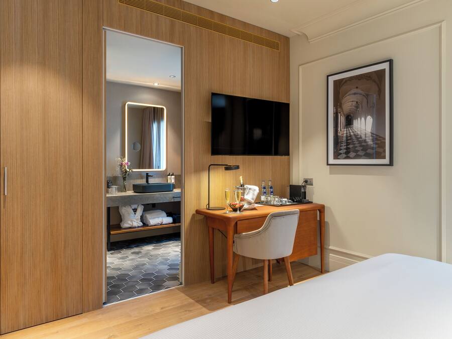 A modern hotel room with a wooden wardrobe, a desk with a chair, a flat-screen TV, a framed picture on the wall, and a bed.