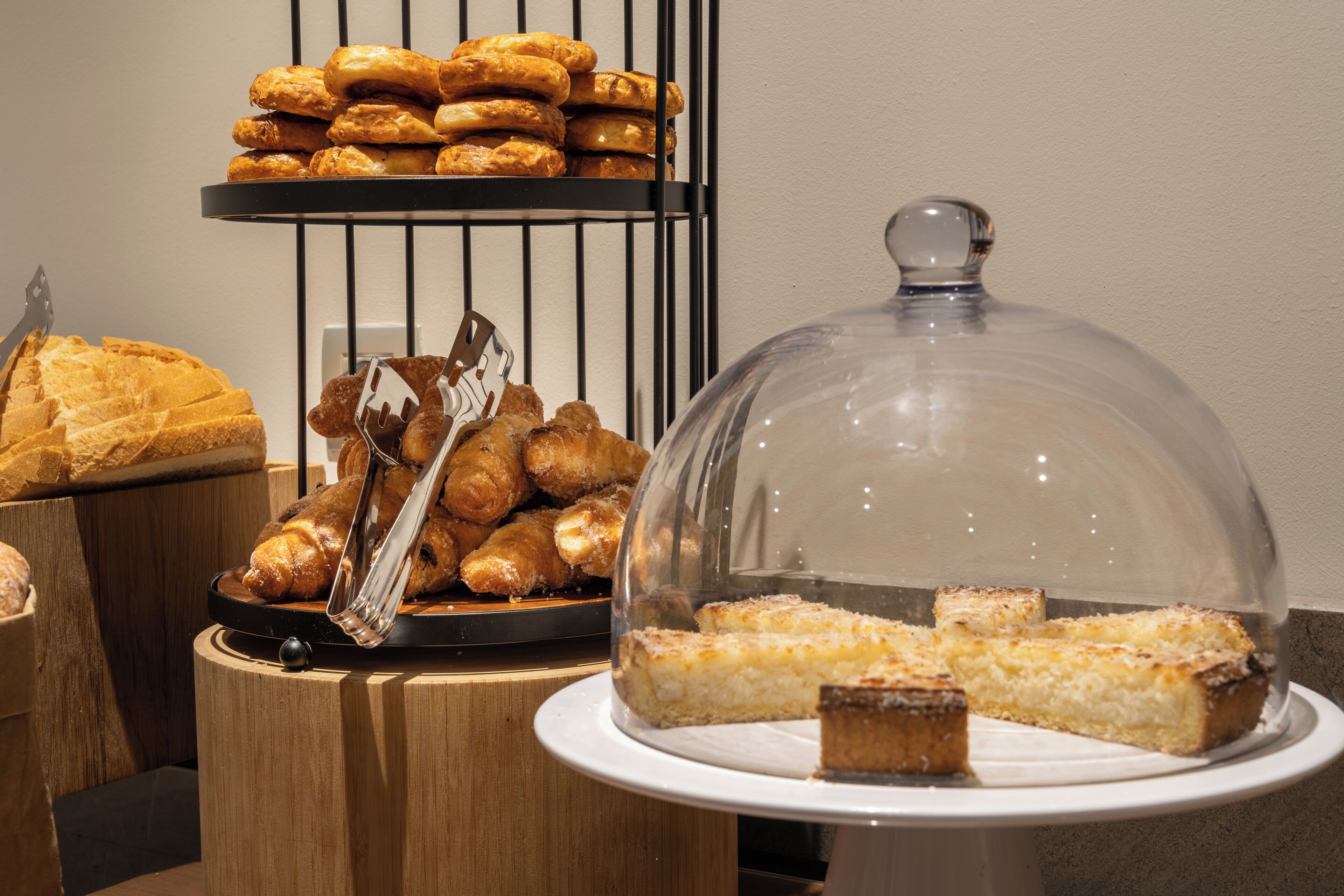 A buffet with various baked goods, including pastries and bread, displayed on wooden stands and a glass-covered dish.