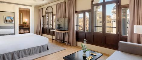 A hotel room with a bed, a desk, a TV, a window with curtains, and a view of buildings.