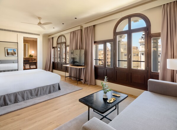 A hotel room with a bed, a desk, a TV, a window with curtains, and a view of buildings. - H10 Palacio Colomera (Córdoba)