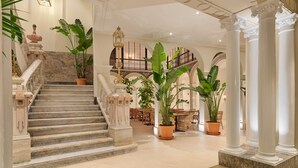 A grand staircase with marble steps and white columns, leading to a dining area with large potted plants and a chandelier. - H10 Palacio Colomera (Córdoba)