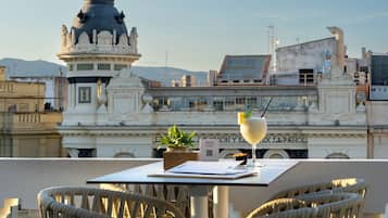 A terrace with white wicker chairs and a table, overlooking a cityscape with historical architecture and a statue.