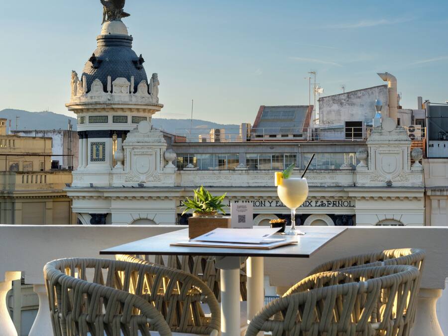 A terrace with white wicker chairs and a table, overlooking a cityscape with historical architecture and a statue.