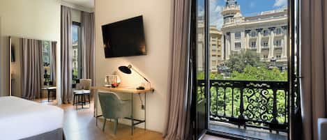 Habitación de hotel con cama, escritorio, silla, televisor y balcón con vista a un edificio.