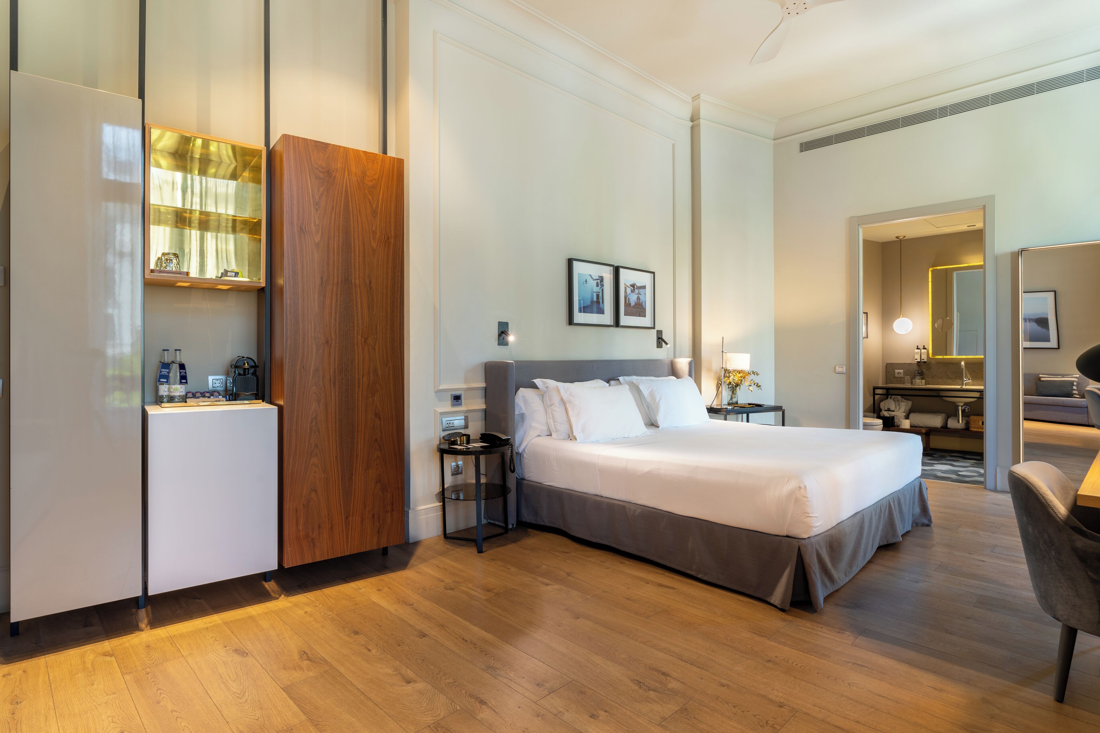 A modern hotel room with a large bed, a wooden wardrobe, a small desk, and a bathroom visible through an open door.