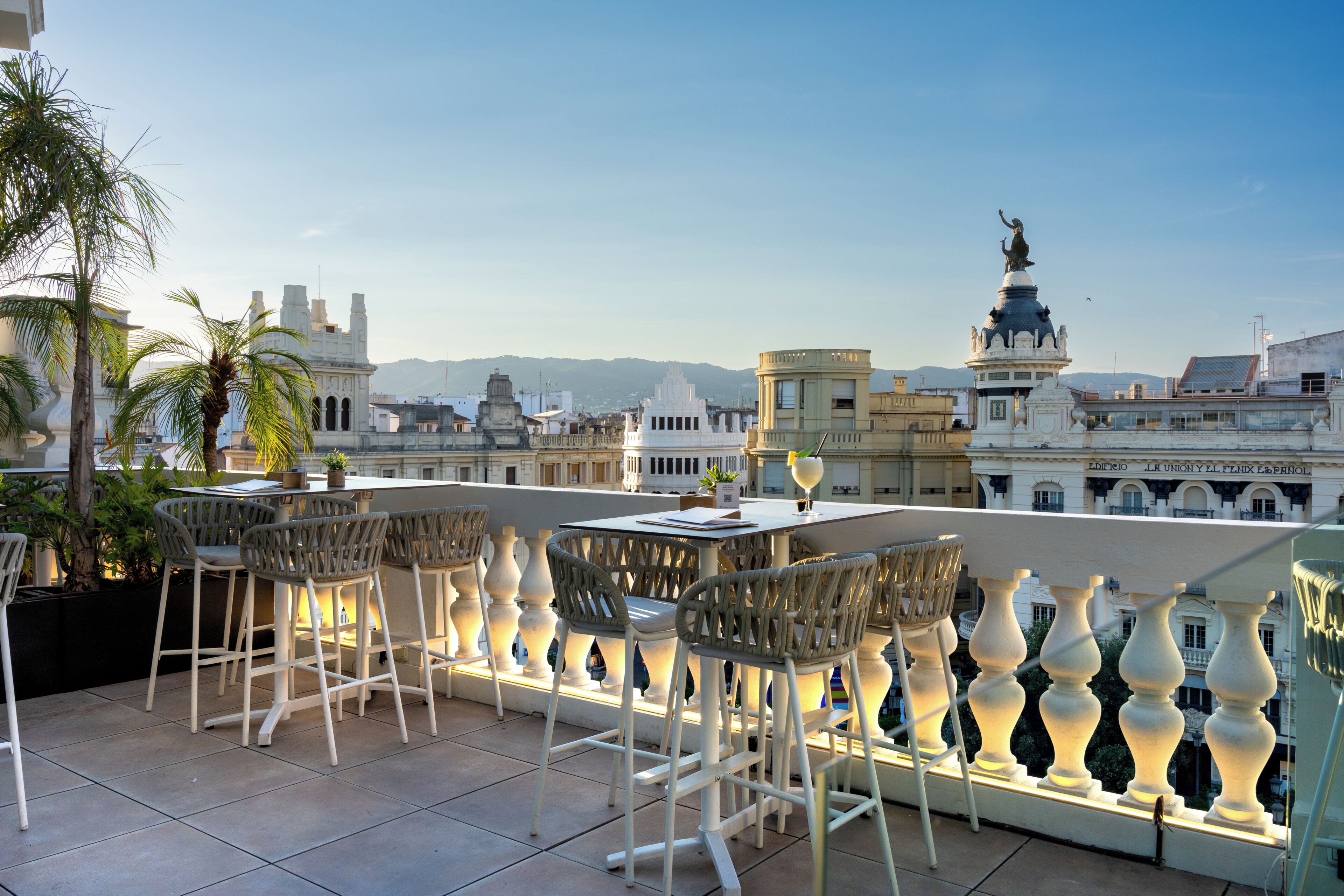 An outdoor seating area with white chairs and tables, overlooking a cityscape with historic buildings and a statue.