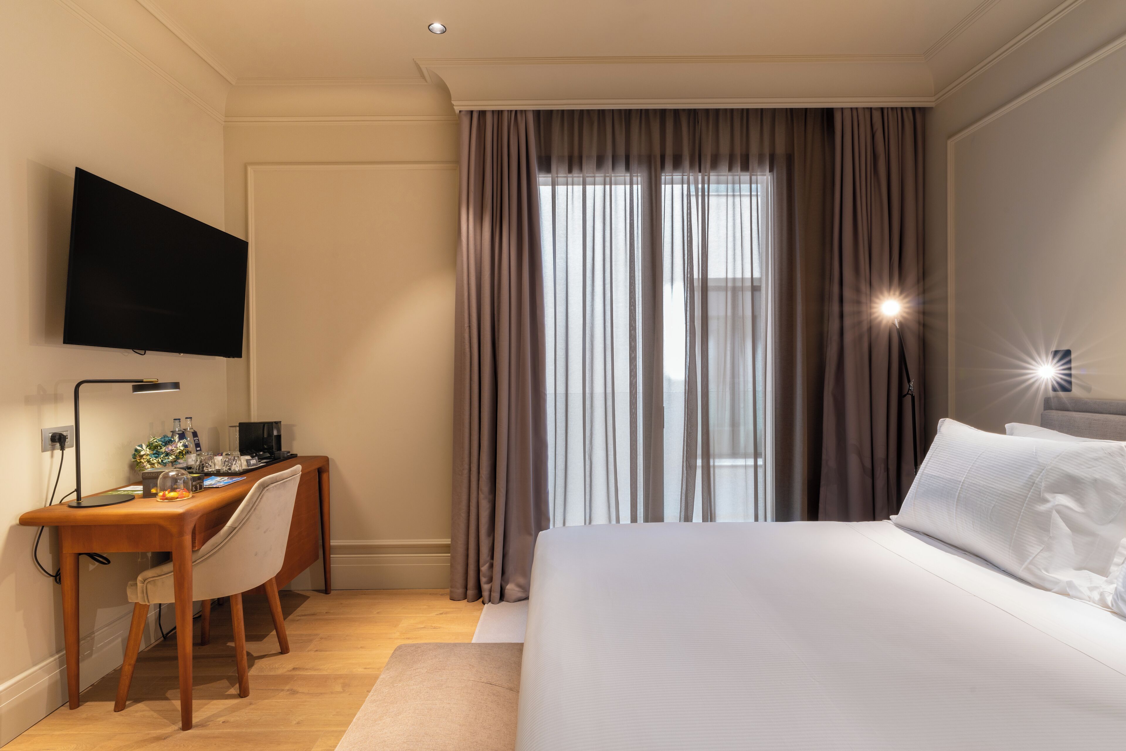 Habitación de hotel con cama, escritorio con silla, televisor de pantalla plana y ventana grande con cortinas.