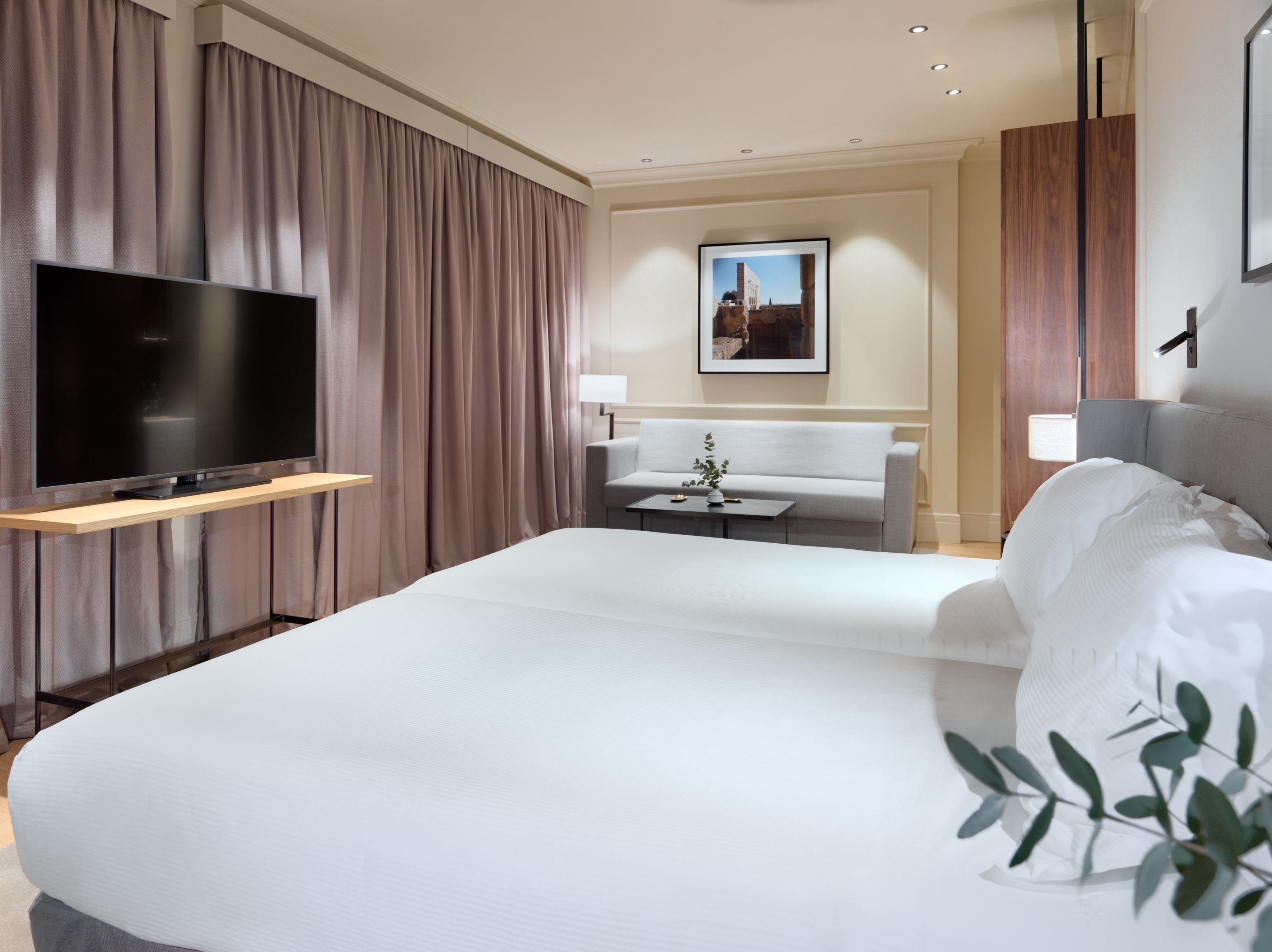 Una habitación de hotel moderna con una cama grande, un televisor de pantalla plana montado en un estante de madera, un sofá gris, una mesita con un florero y un cuadro en la pared.