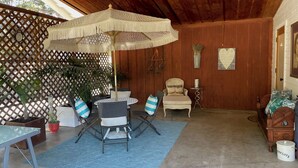 Un patio couvert avec une table ronde, des chaises et un parasol suspendu. On y trouve des plantes en pot, une chaise en bois et un lit recouvert d’une couverture bleue.
