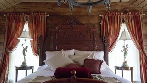 Una cama con estructura de madera y un cabecero decorativo, dos mesitas de noche con lámparas y cortinas rojas.