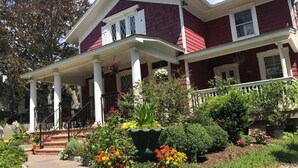 Una casa de dos pisos con un porche, columnas y un jardín con diversas plantas y flores.