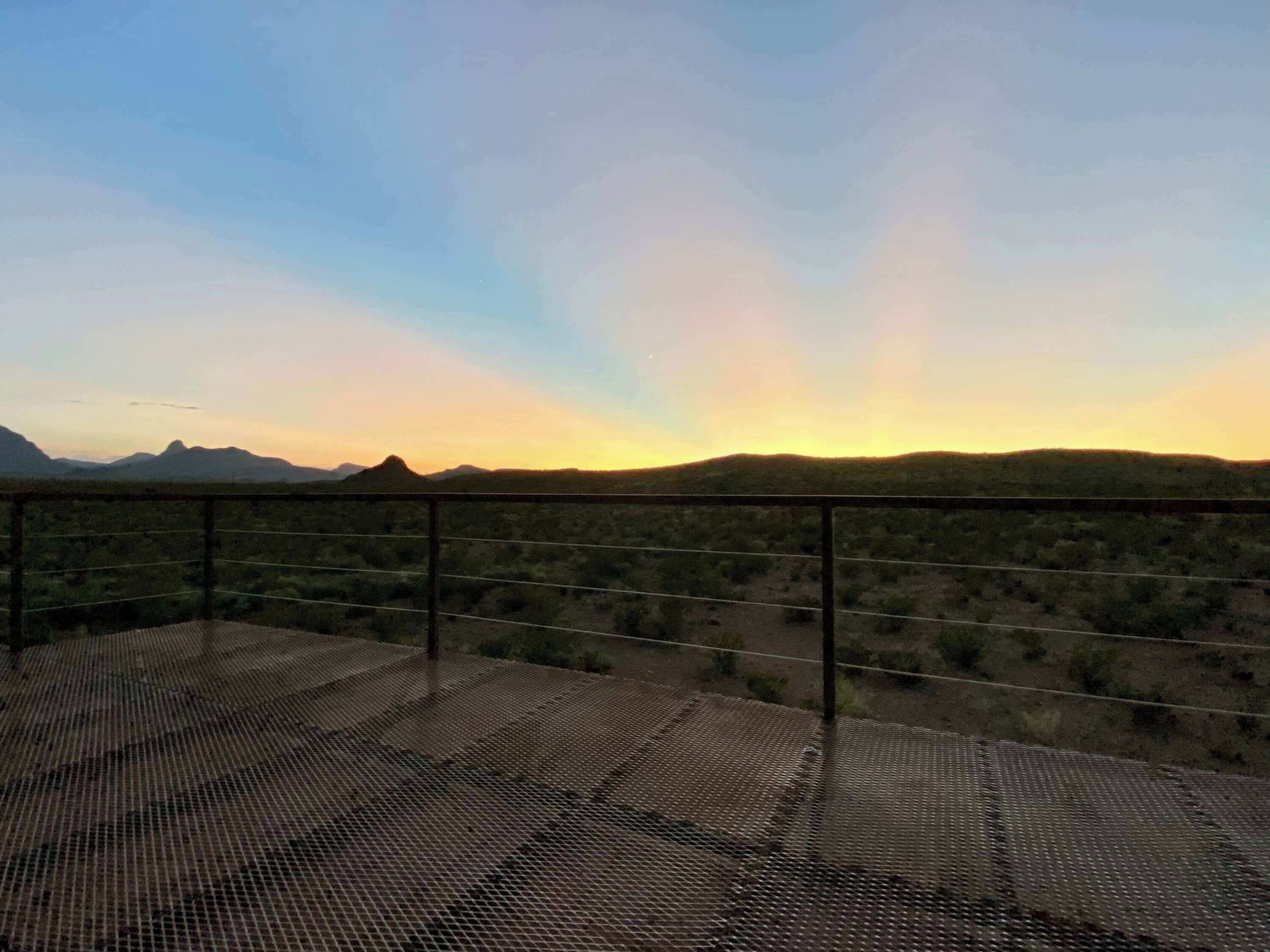A balcony with a metal grid floor overlooking a desert landscape with distant mountains.