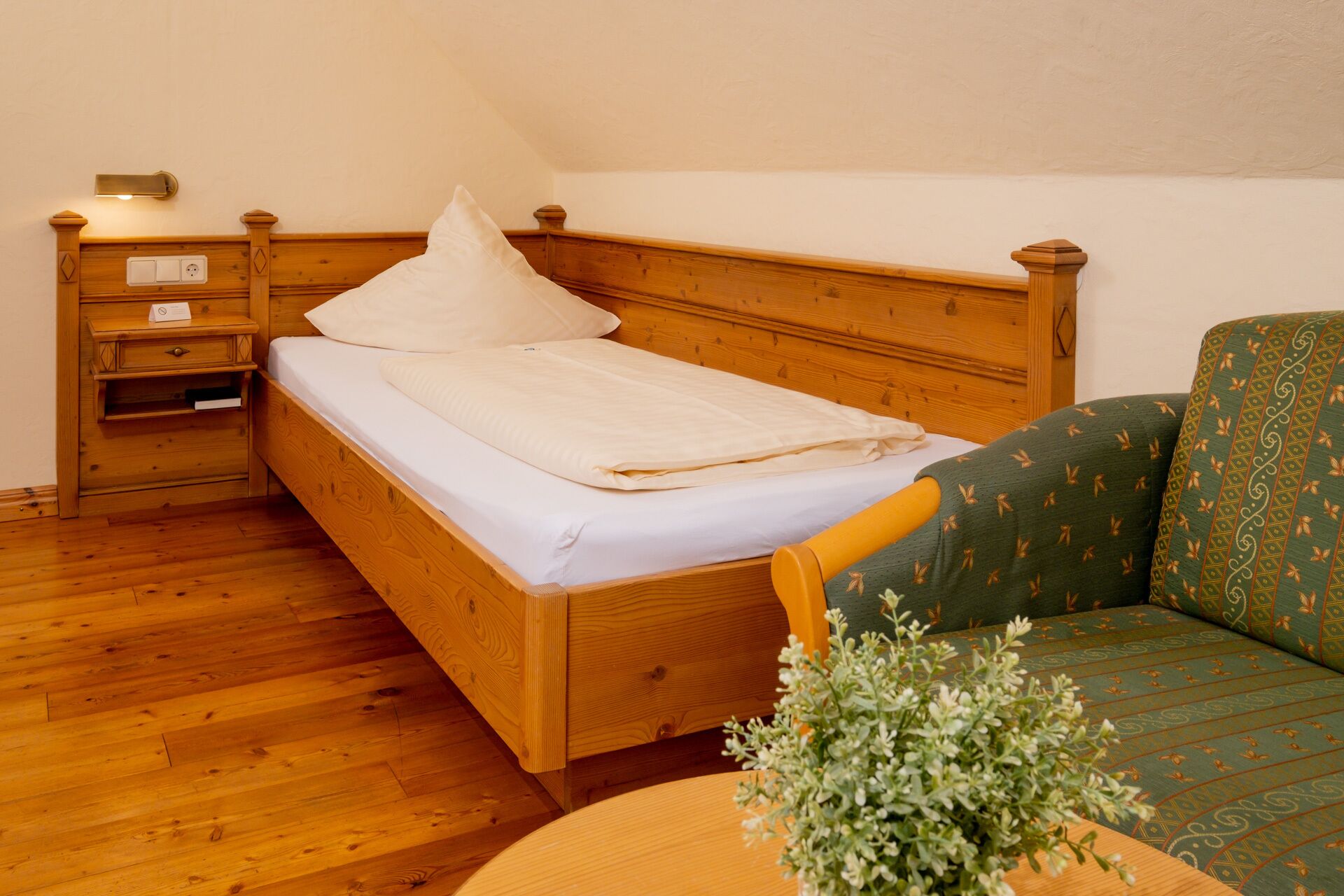 Een houten bed met matras en kussens, een nachtkastje met lamp, en een groene sofa met een patroon.