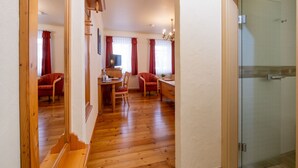 Un pasillo con piso de madera que conduce a un comedor con dos sillas y una mesa, un televisor sobre un mueble bajo, y una puerta de cristal a la derecha.