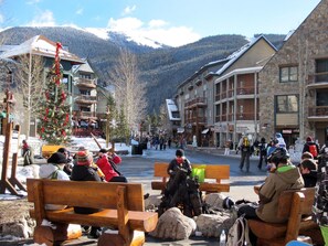 A snowy outdoor area with benches, a Christmas tree, and buildings in the background. - Top floor condo, mountain views, steps to village, free shuttle, 1 min to slopes (Keystone)