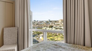 A hotel room with a large window, a bed with a floral bedspread, a chair, and curtains. - Amazing Beachfront Condo in the heart of Destin - w/FREE Beach Setup March-Oct (Destin)